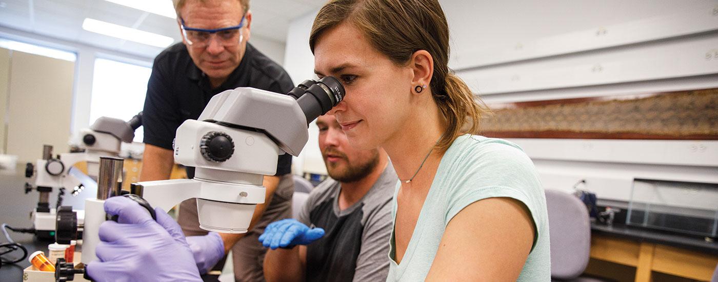 A Frostburg State student looks into a microscope while a professor gives an explanation and another student looks on